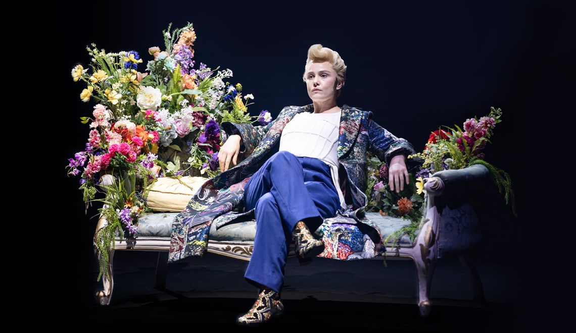 A young blonde woman with short hair sits on an ornate chaise lounge, with a large bouquet of flowers by her right hand side. She is wearing a white top with blue pants, and an ornate, flowing silk gown over the top.