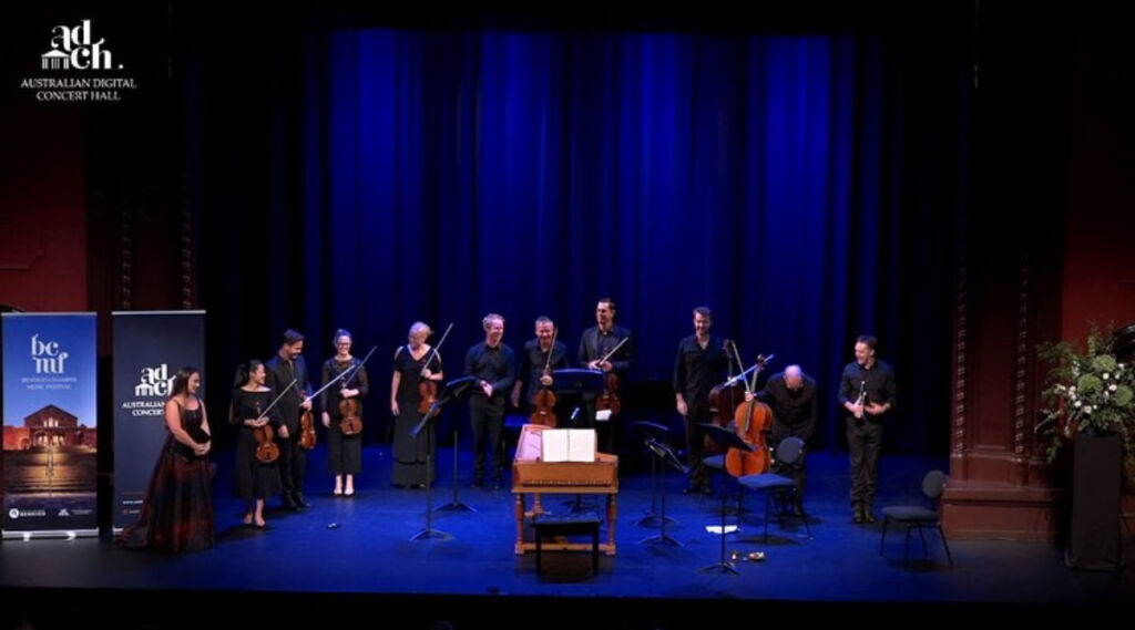 Eleven musicians stand in a line on a stage at the end of a performance. A light brown wooden harpsichord sits in the middle of the stage.
