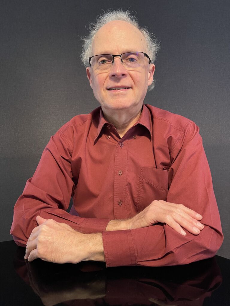 Australian composer Colin Spiers. An older white man with wispy grey hair, spectacles, and wearing a red button-up shirt.