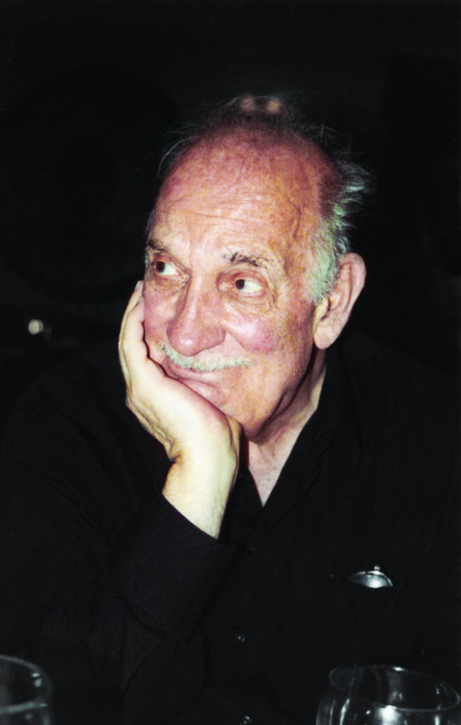 A photo of American composer George Crumb. An older man, mostly bald, with a small amount of grey hair at the back of his head, and a grey moustache. He is resting his chin on his right hand. He is wearing a black shirt.
